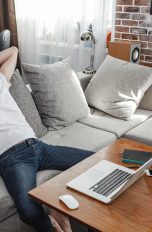 Eine Person sitzt auf einem Sofa. Vor ihr auf dem Tisch ist ein MacBook zu sehen, neben dem Sofa ein Lautsprecher.