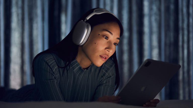 Apple One Eine Person liegt auf einem Bett, hat Apple AirPods Max auf und schaut auf ihr iPad.