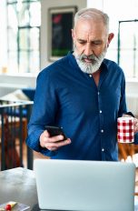 Eine Person steht mit einer Tasse in der Hand vor einem Laptop. In der Hand hält sie ein iPhone.