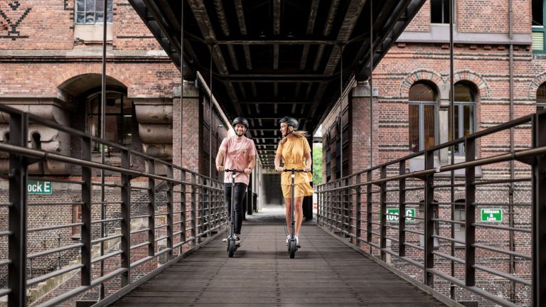 Ninebot KickScooter Max G30D II Zwei Personen fahren mit zwei KickScooter Max G30D II über eine Brücke in der Hamburger Speicherstadt.