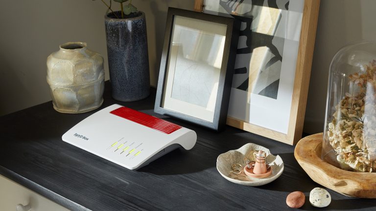 Eine Fritzbox 7590 AX steht auf einer schwarzen Kommode neben Deko-Artikeln.