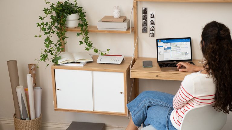 FRITZBox 7590 Eine Frau sitzt an einem Schreibtisch vor ihrem Laptop. Darauf zu sehen ist die Benutzeroberfläche einer Fritzbox. Diese steht neben ihr auf einem kleinen Schrank.