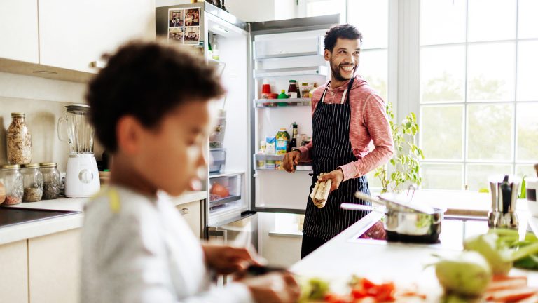Eine Person holt etwas aus dem Kühlschrank. Im Vordergrund ist ein Kind zu sehen, dass Gemüse schneidet.