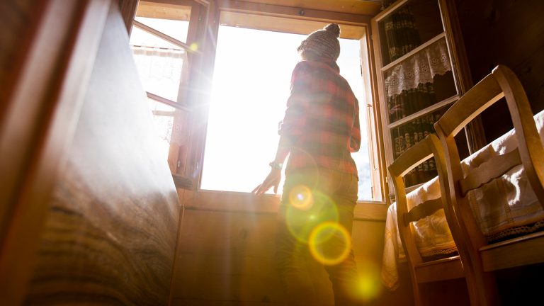 Lens Flare Eine Person steht an einem Fenster und schaut hinaus. Die Sonne erzeugt Blendenflecken im Bild.