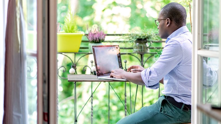 Outdoor-Homeoffice Eine Person sitzt mit einem Laptop auf dem Balkon. Das Display spiegelt.