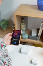 Eine Person steht vor einem Tisch, auf dem ein Bewegungsmelder von Philips Hue, die Philips Hue Bridge und eine Lampe stehen. Sie schaut auf ihr Smartphone, das den Startbildschirm der Philips Hue App zeigt.