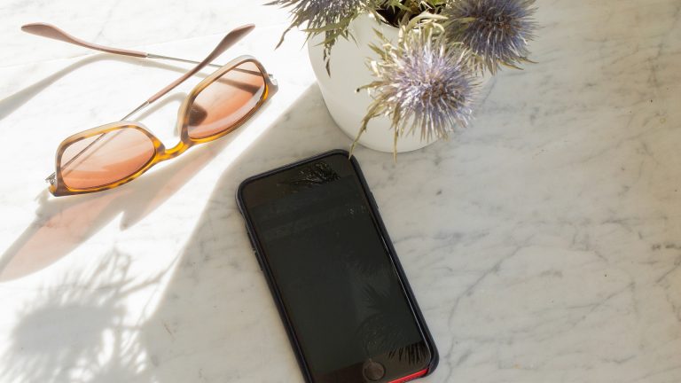 Ein Smartphone liegt auf einem Marmortisch im Schatten. Daneben liegt eine Sonnenbrille.