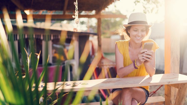 Smartphone im Sommer Eine Frau sitzt auf einer Holzbank am Strand und schaut auf ihr Handy.