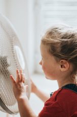 Ein Kind steht vor einem eingeschalteten Ventilator, seine Haare wehen im Wind.