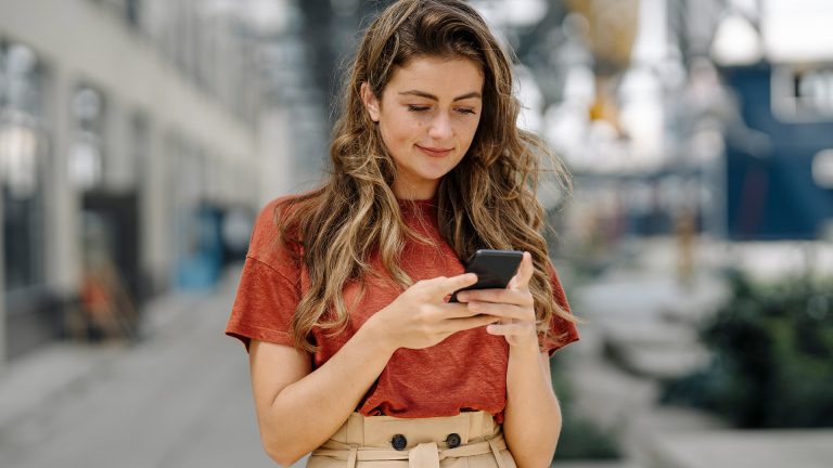 WhatsApp Broadcast Eine junge Frau steht in einer Einkaufspassage und schaut auf ihr Smartphone.