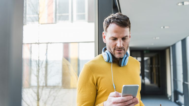 Ein Mann steht in einem Flur eines Bürogebäudes und schaut auf sein Smartphone. Um seinen Hals baumelt ein Paar Kopfhörer.