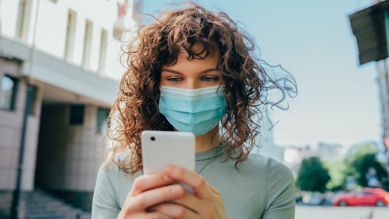Eine Frau mit Gesichtsmaske schaut auf ihr Smartphone. Sie ist der Kamera zugewandt, das Smartphone selbst ist nur von hinten zu sehen.