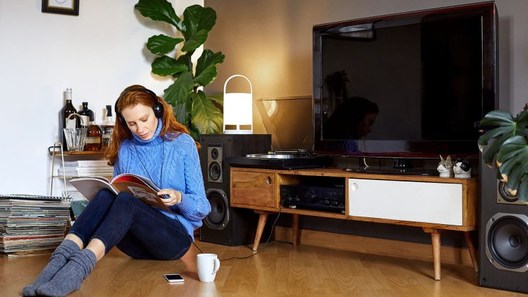 Eine Frau sitzt auf dem Fußboden und liest in einem Magazin, hinter ihr sind ein Sideboard mit TV-Gerät und Lautsprechern rechts und links zu sehen.