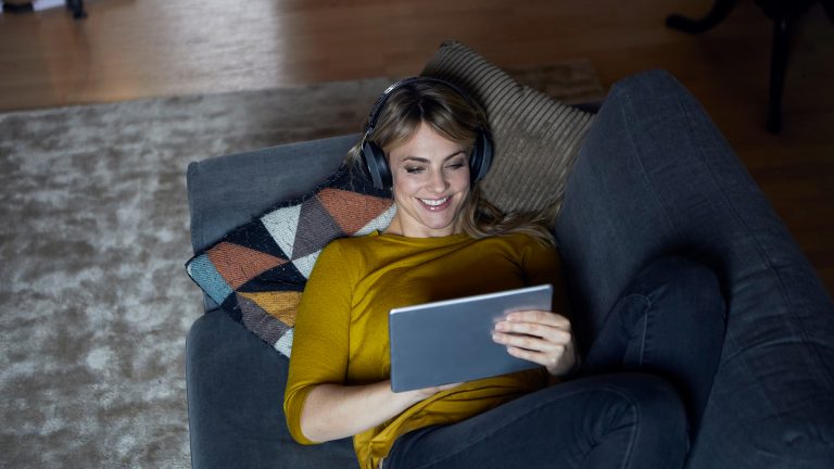 Online-Spiele Eine Person liegt auf dem Sofa, trägt Kopfhörer und spielt etwas auf einem Tablet.