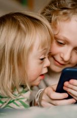 Zwei Kinder liegen auf dem Bett und schauen auf ein Smartphone.