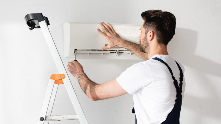 Eine Person auf einer Leiter schraubt die Abdeckung einer Split-Klimaanlage auf.