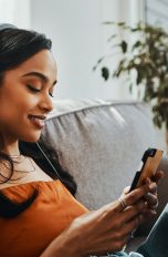 Eine junge Frau sitzt auf dem Sofa, schaut auf ihr Smartphone und hört über einen Kopfhörer Musik.