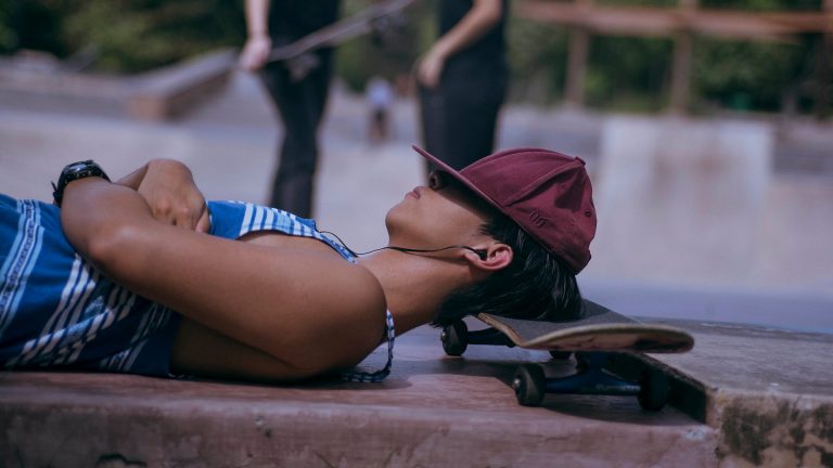Musik Streaming Spotify Eine junge Frau liegt mit dem Kopf auf einem Skateboard, die Baseball-Kappe ins Gesicht gezogen, im Ohr die Kopfhörer um Musik zu streamen.