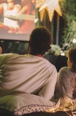 Ein Paar liegt auf Kissen und Decken vor einer Leinwand und schaut einen Film im eigenen Garten.