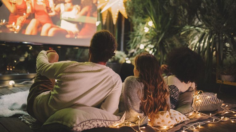 Ein Paar liegt auf Kissen und Decken vor einer Leinwand und schaut einen Film im eigenen Garten.