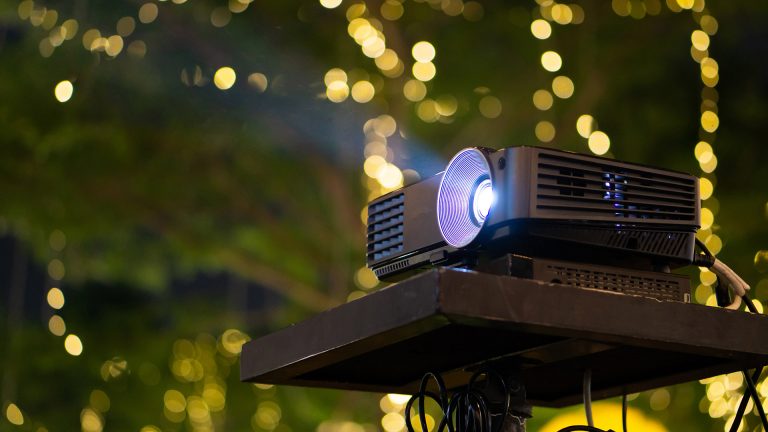 Outdoor Kino Auf einem kleinen Tisch steht ein Beamer vor einer verschwommenen, illuminierten Gartenkulisse.