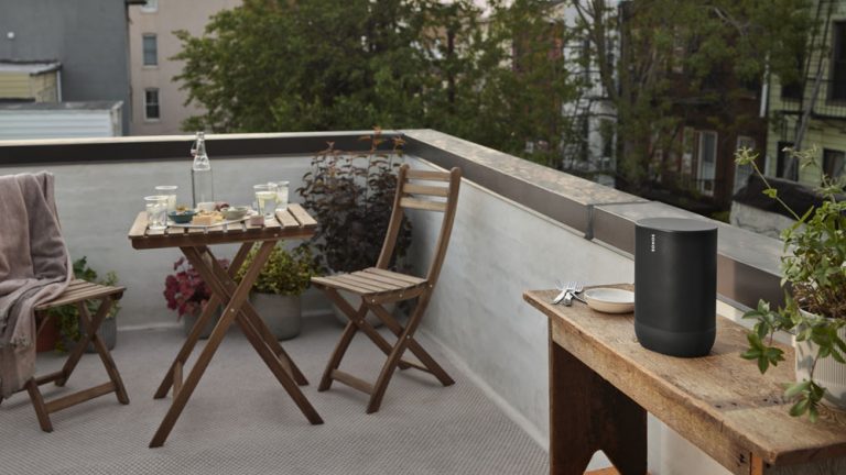 Outdoor Kino Auf einem Balkon steht ein Bluetooth-Lautsprecher auf einem Tisch, im Hintergrund ein kleiner gedeckter Tisch für Zwei.