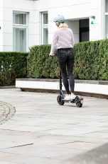 Eine Person fährt auf einem E-Scooter vor einer großen Hecke.