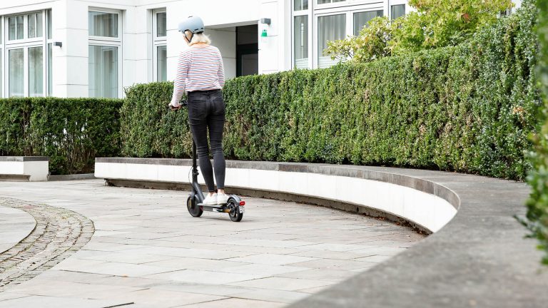 Eine Person fährt auf einem E-Scooter vor einer großen Hecke.