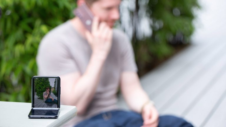 Samsung Galaxy Z Flip2 Im Vordergrund steht ein halb aufgeklapptes Samsung Galaxy Z Flip auf einem Tisch. Im Hintergrund ist unscharf eine Person zu erkennen, die gerade telefoniert.