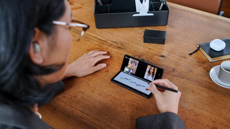 Samsung Galaxy Z Fold3 Eine Person sitzt vor einem halb aufgeklappten Samsung Galaxy Z Fold3 5G und schreibt mit einem S Pen auf der unteren Bildschirmhälfte, während die obere ein Videotelefonat zeigt.
