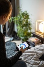 Eine Person sitzt auf einem Bett und steuert per Smartphone eine Lampe vor ihr.