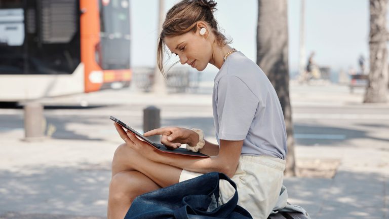 JBL Tune 130NC Eine Person sitzt auf einer Bank und bedient ein Tablet. Im Ohr hat sie die JBL Tune 130NC.