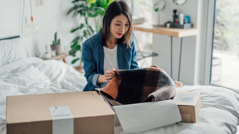 Eine Person sitzt mit einem geöffneten Paket auf dem Bett und hält eine bestellte Ware in der Hand. Eine Person sitzt mit einem geöffneten Paket auf dem Bett und hält eine bestellte Ware in der Hand.