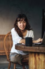 Eine Person sitzt an einem Tisch, vor ihr steht ein aufgeklappter Laptop. Sie spricht zu einem smarten Lautsprecher.