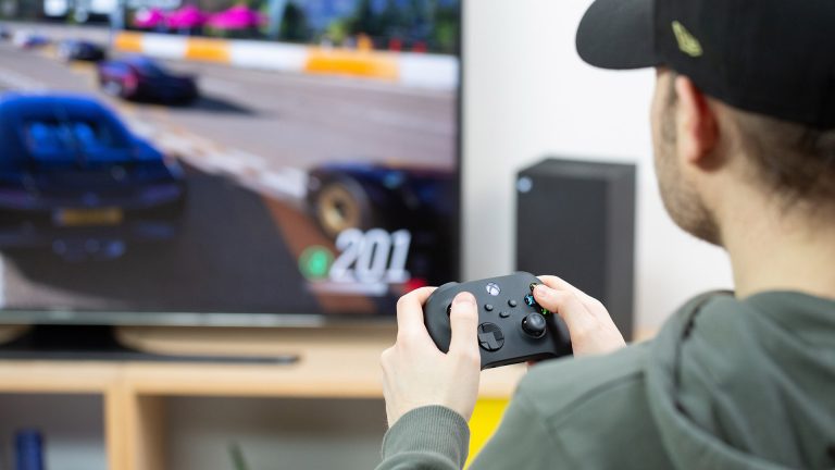 Microsoft Xbox Eine Person hält den Controller der Xbox Series X in der Hand. Die Konsole ist im Hintergrund zu sehen.