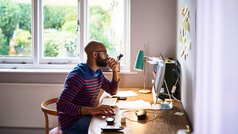 NAS Eine Person sitzt an einem Schreibtisch und schaut auf einen iMac. Neben Maus und Tastatur liegen weitere Arbeitsutensilien auf dem Tisch, unter anderem ein Telefon und mehrere Dokumente.