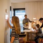 Eine junge Person bedient ein Smarthome-Element an einer Wand. Im Hintergrund ist eine Familie zu sehen, die sich um einen Esstisch versammelt.