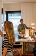 Eine junge Person bedient ein Smarthome-Element an einer Wand. Im Hintergrund ist eine Familie zu sehen, die sich um einen Esstisch versammelt.