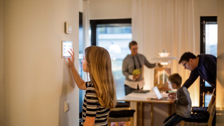 Eine junge Person bedient ein Smarthome-Element an einer Wand. Im Hintergrund ist eine Familie zu sehen, die sich um einen Esstisch versammelt.