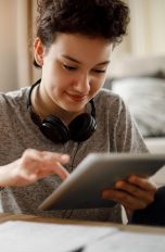 Eine Person sitzt an einem Tisch. In der Hand hält sie ein iPad, das sie mit der anderen bedient.