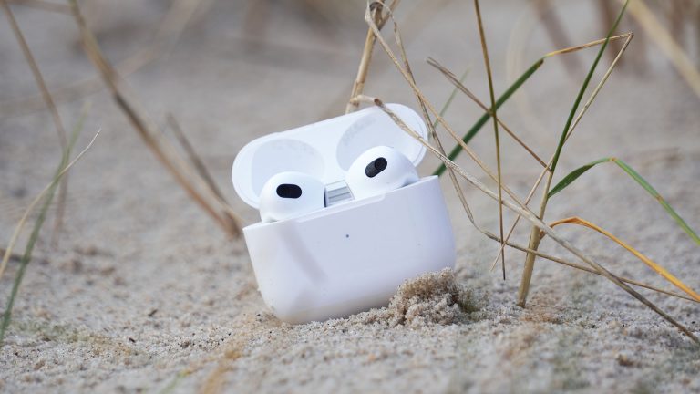 Apple AirPods (3. Gen.) AirPods der dritten Generation stehen in ihrem Ladecase im Sand.