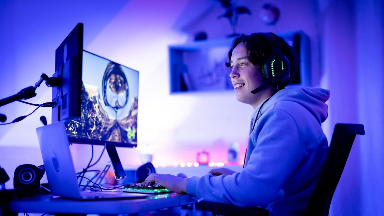 Eine Person spielt an einem Tisch, auf dem ein Laptop und zwei Monitore stehen.