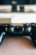 Ein DualShock-4-Controller) liegt auf einer Sofalehne. Im Hintergrund ist ein Fernseher zu erkennen.