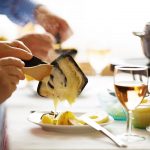 Eine Person schiebt Kartoffeln und Käse mit einem Holzlöffel aus einer Raclettepfanne.
