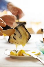 Eine Person schiebt Kartoffeln und Käse mit einem Holzlöffel aus einer Raclettepfanne.
