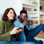 Zwei Personen hocken vor einem Sofa. Beide halten einen PlayStation-4-Controller in den Händen.
