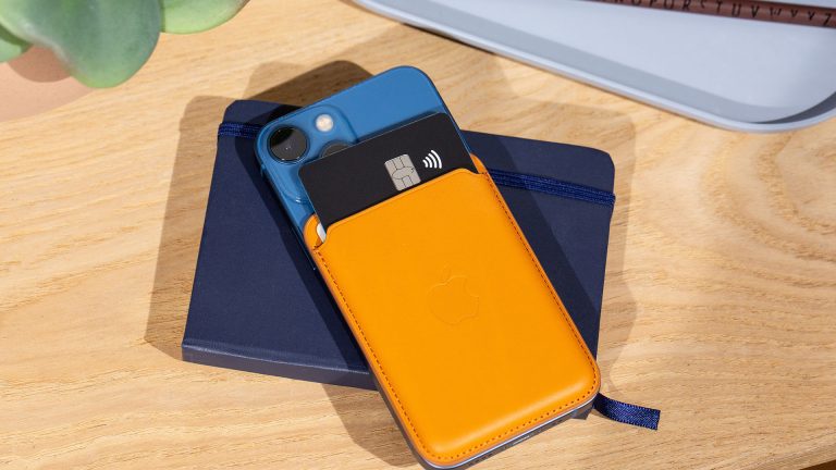 Geschenkideen für Apple-Fans Auf einem Holztisch liegt ein iPhone 12 mini dekorativ auf einem blauen Notizbuch. Die Rückseite des iPhones ziert ein hellbraunes Leder Wallet.