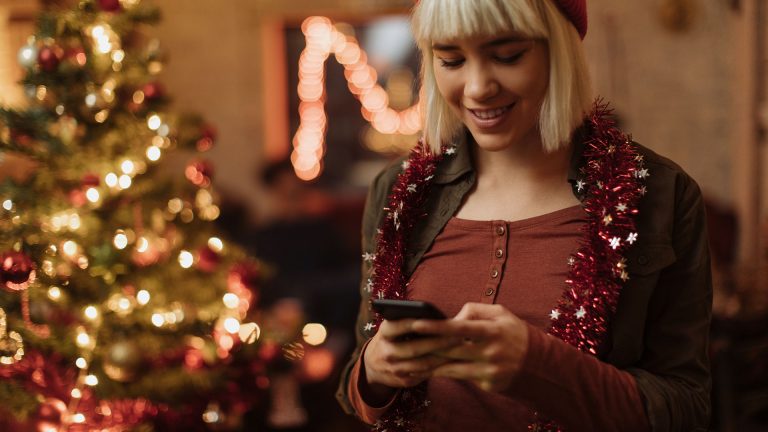 Smarte Lichterkette für den Weihnachtsbaum Eine junge Frau steht mit Ihrem Smartphone vor einem mit Lichterketten geschmückten Weihnachtsbaum.