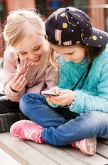 Zwei Kinder sitzen auf einer Bank im Freien und schauen gemeinsam auf ein Smartphone.