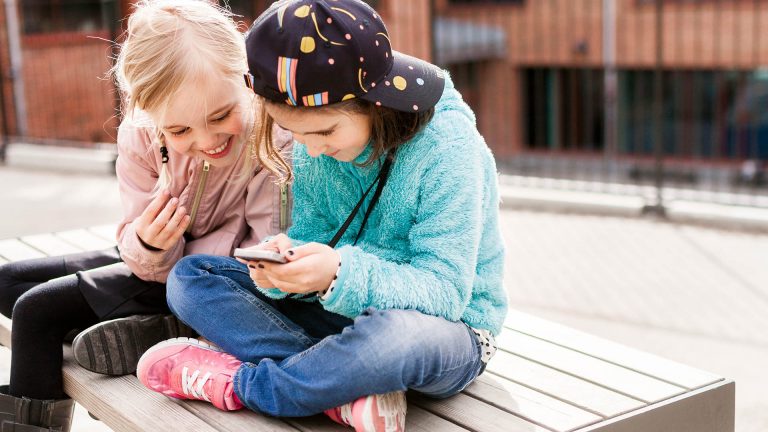 Zwei Kinder sitzen auf einer Bank im Freien und schauen gemeinsam auf ein Smartphone.